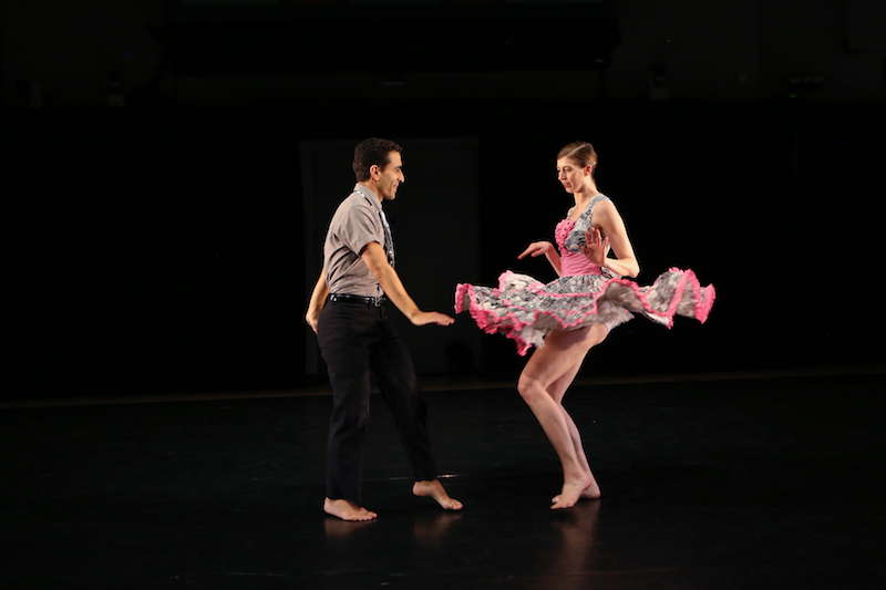 Two dancers do the twist facing one another. Their hands are flexed and they're barefoot. The woman's pink skirt cascades around her as she twists.